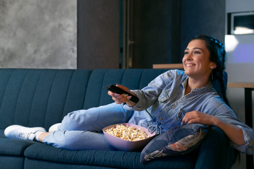 Mujer recostada con palomitas y mando a distancia viendo un televisor